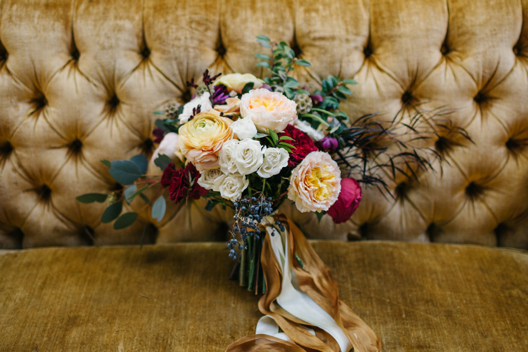 Lush bouquet of burgundy, peahc, and gold peonies and garden roses sitting on a vintage mustard couch for outdoor Orlando garden wedding