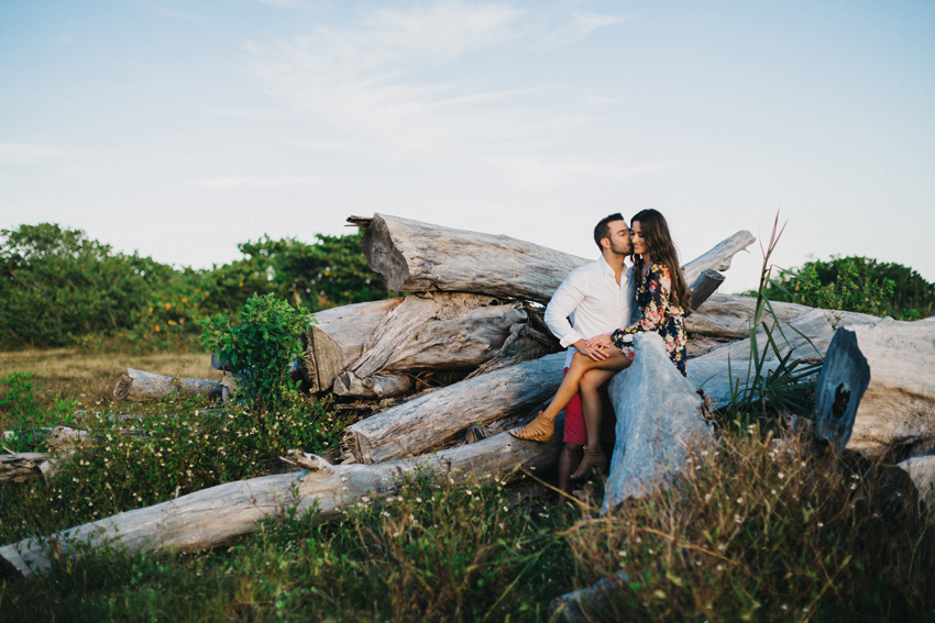 Adventure engagement session at sunset in a field with the couple sitting on a pile of old wooden logs