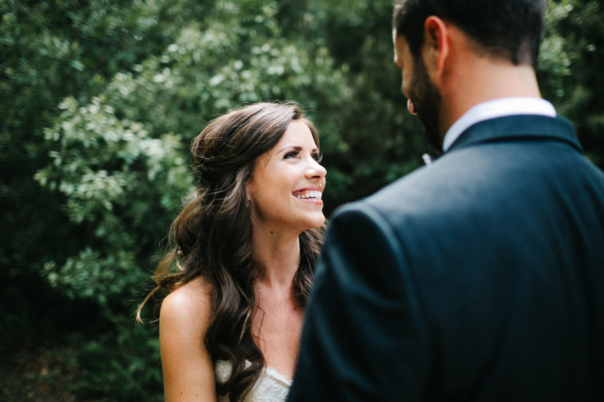Beautiful bride with her hair half up smiling up at her groom during their first look in the woods