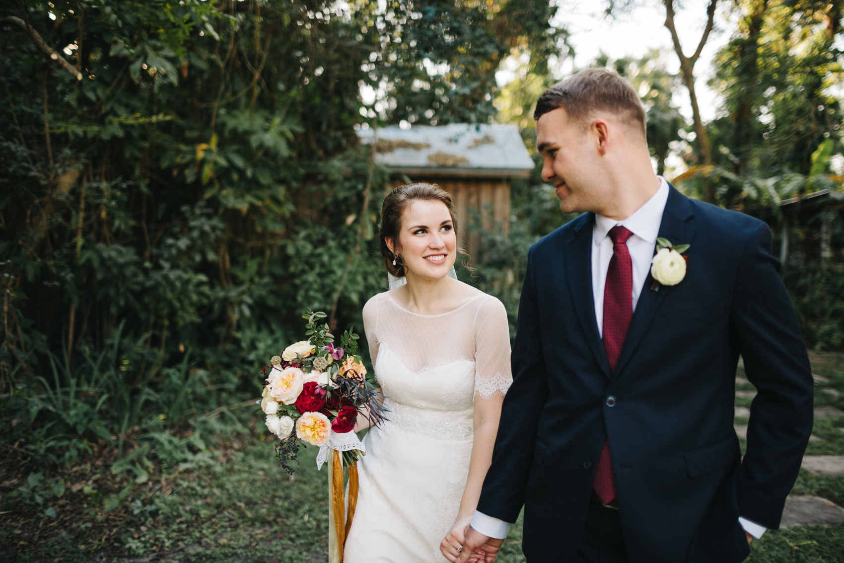 bride smiling at her groom carrying a lush burgundy, peach, and gold wedding bouquet