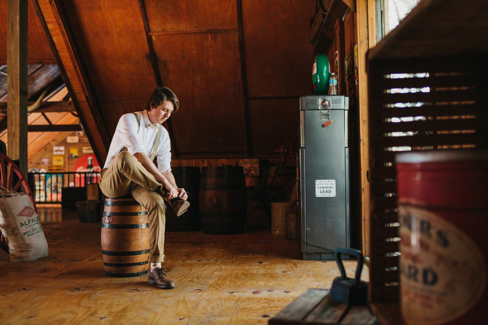 groom wearing suspenders and khaki pants putting on his shoes inside the rustic barn