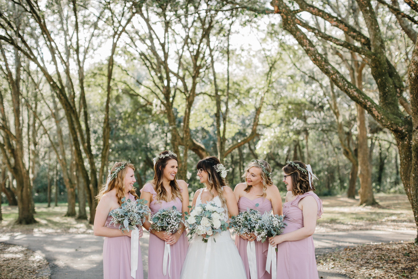 bridesmaids wearing dusty pink gowns laughing with the bride under the oak trees at the Lange Farm barn venue