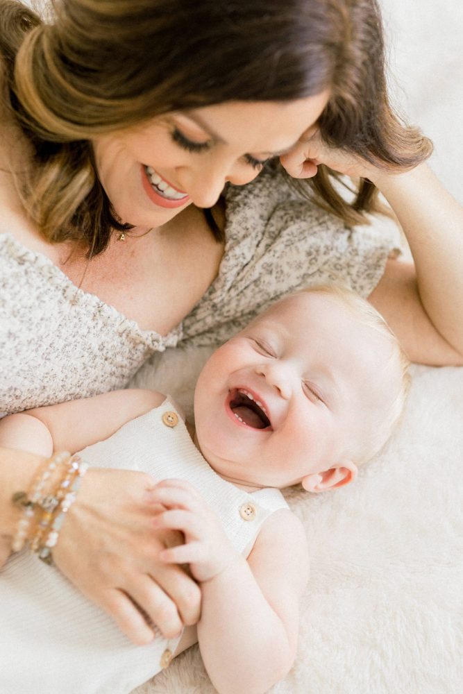 Photoshoot of Mom and baby laughing and snuggling on the bed in an all white studio in south tampa